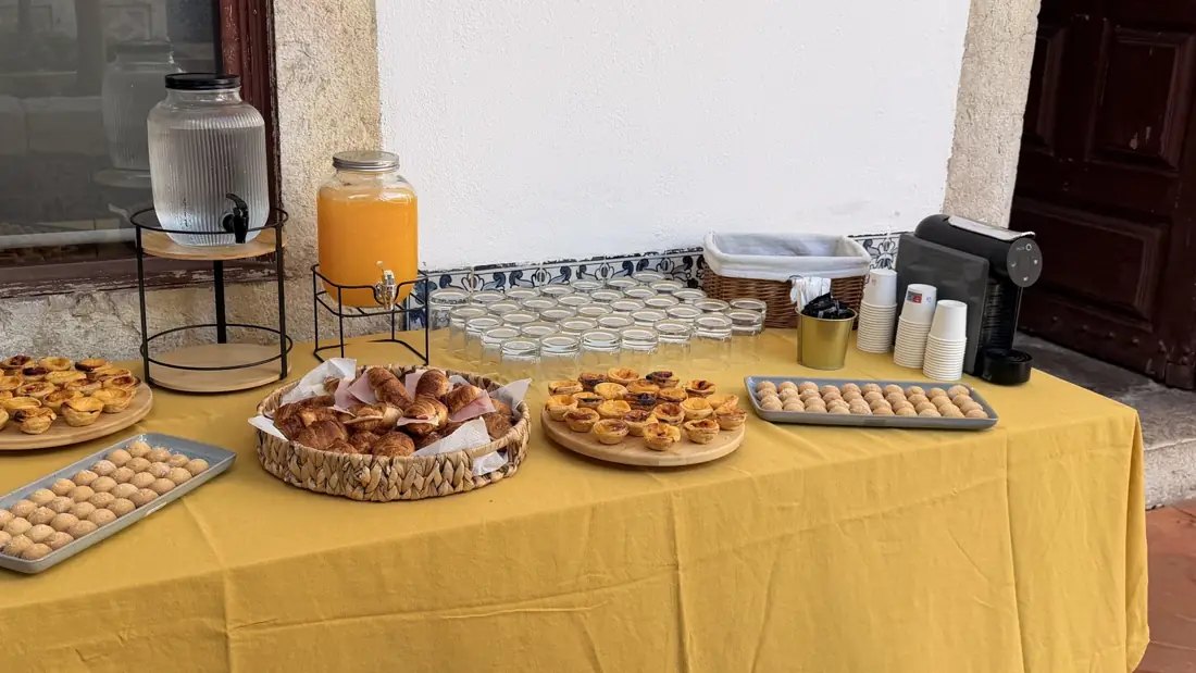 Mesa de coffee break com pastéis de nata, croissants, sumo e café junto a azulejos